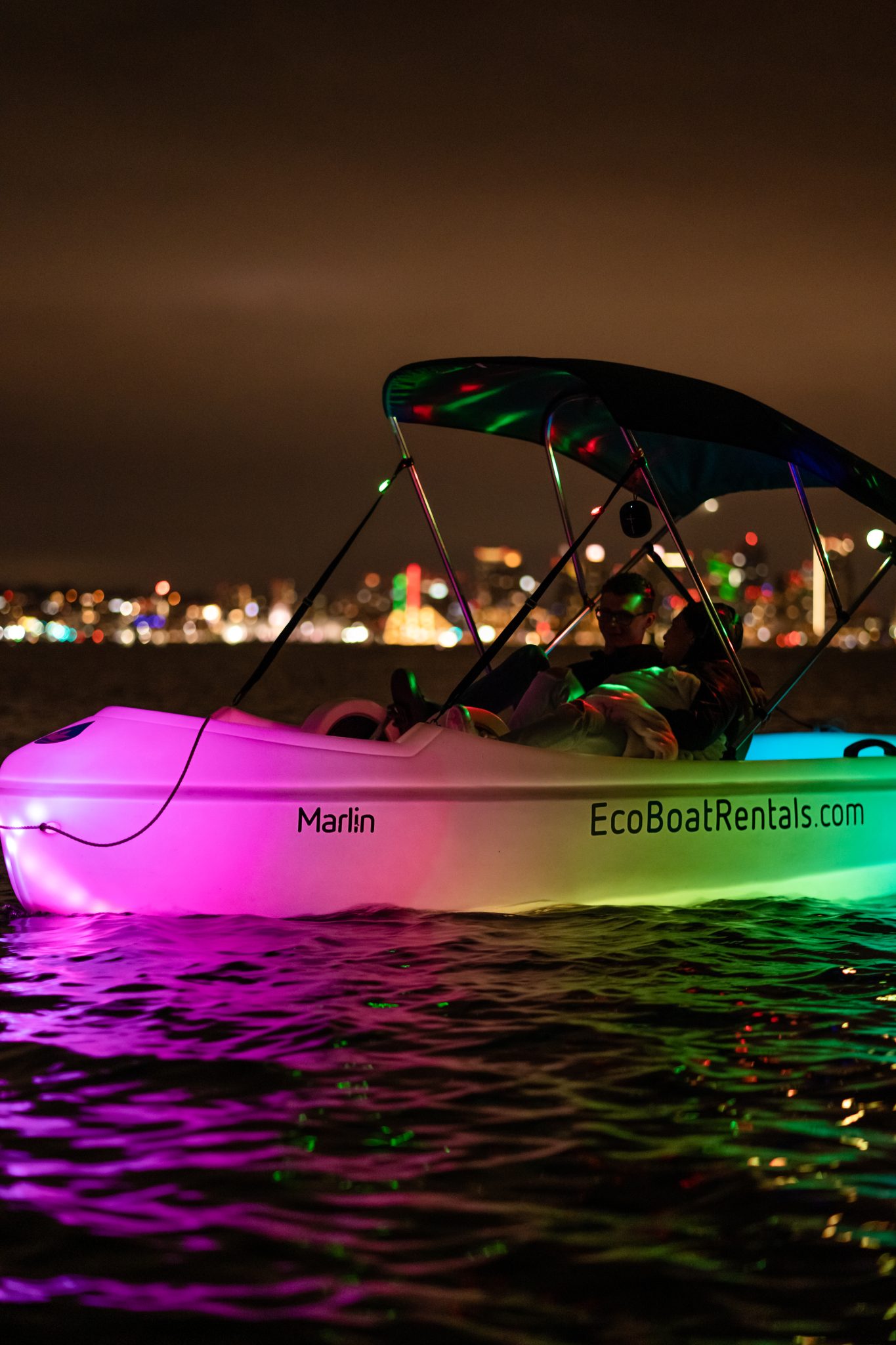 Nighttime boat with a colorful LED underglow on the water, city lights in the distance and a canopy overhead on the boat.