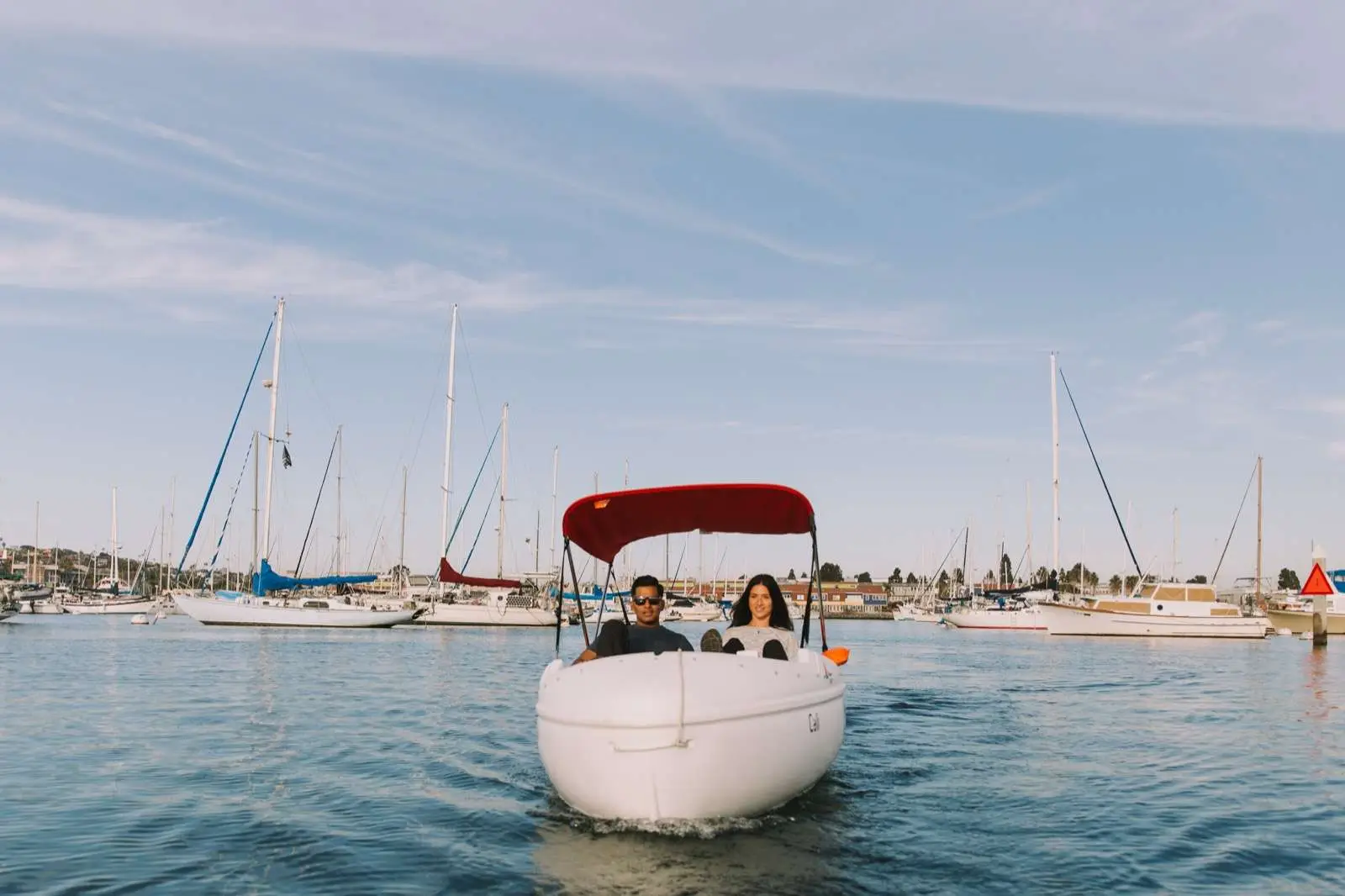 Two people ride a small white motorboat with a red canopy across a calm marina, sailboats in the background.